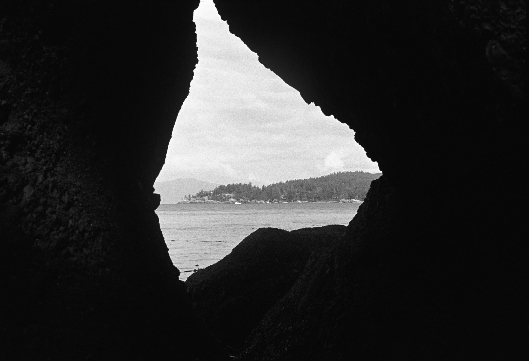 Looking out through an opening in a sea cave. The rock is almost entirely black; the gap frames open water, a far wooded shoreline with a few houses, and a single cloud.