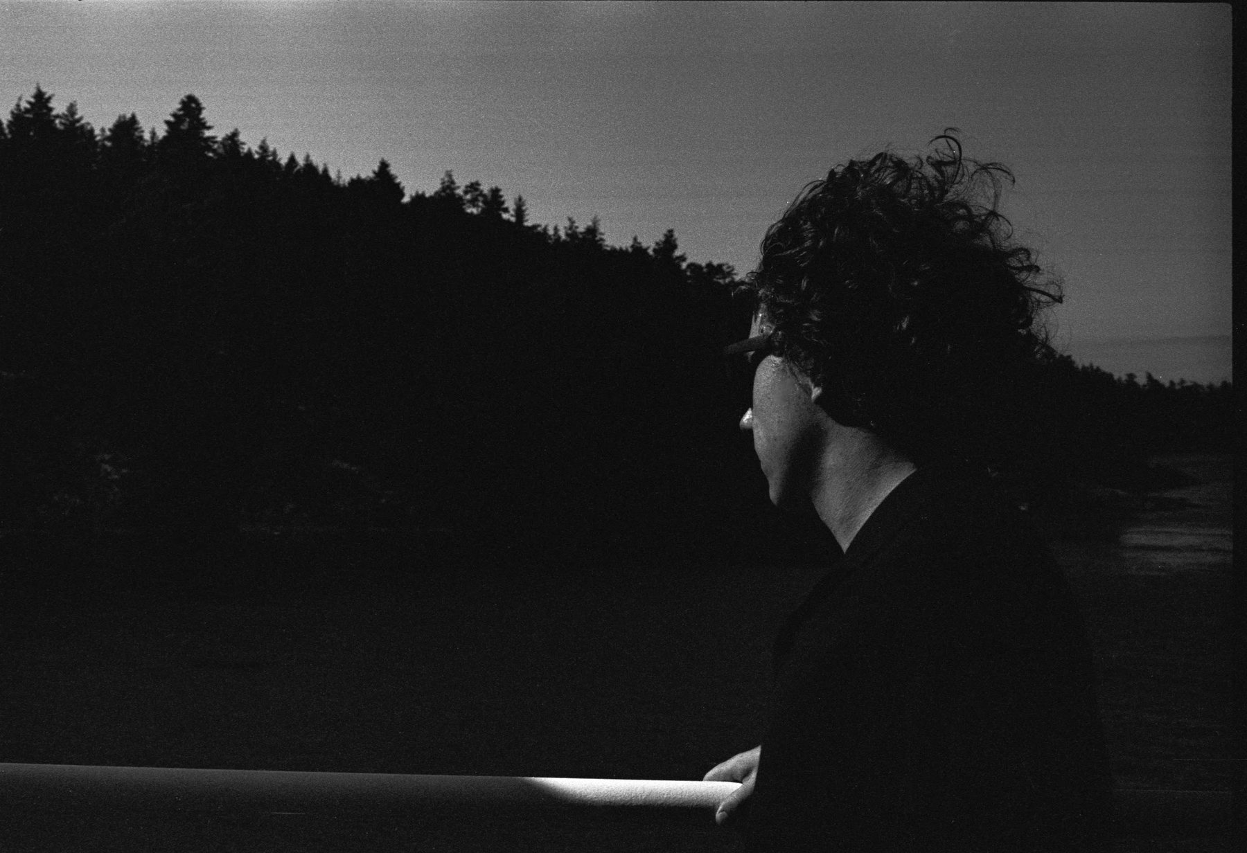 A person in a cap seen in profile at dusk, hair lit from behind, resting against a boat railing with a dark treeline behind them.