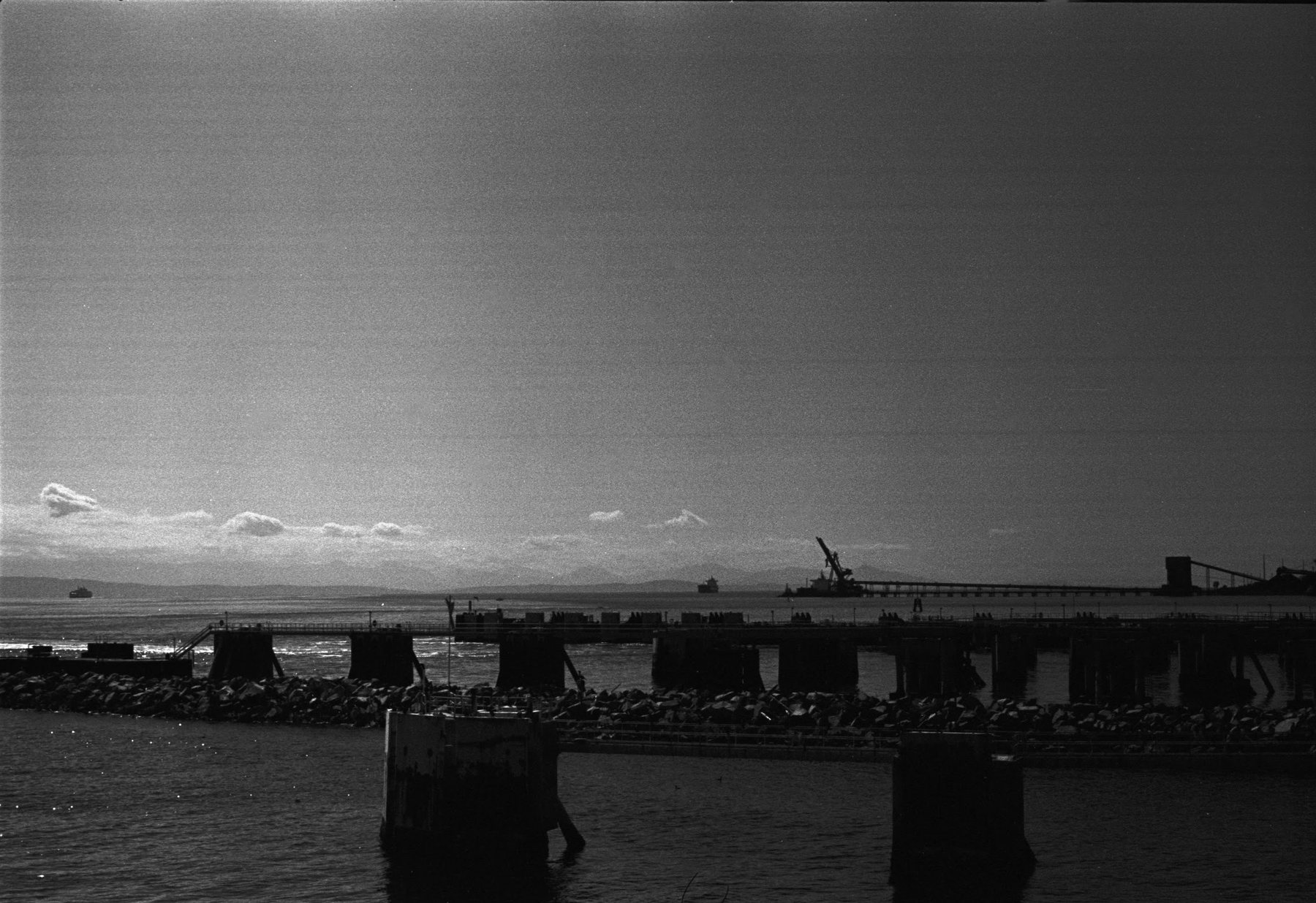 A long pier on pilings running out into the water, with industrial cranes on the far shore under a wide sky.